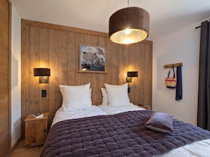 A bedroom with a wooden headboard, two bedside lamps, a large bed with a dark quilt, a wall-mounted picture, and a small bedside table with a book. - Apartment with village and mountain view - 6 guests (Notre-Dame-de-Bellecombe)
