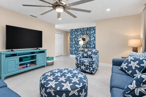Ein Wohnzimmer mit Flachbildfernseher, einem turquoisefarbenen Unterhaltungsmöbel, einem osmanischen Hocker mit Sternenmuster, einem blauen Sofa und einem Deckenventilator.