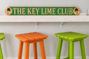 Een houten bord met de tekst 'THE KEY LIME CLUB' en twee krukken, een oranje en een groene, voor een lichtgekleurde muur.