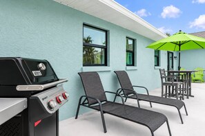 Een terras buiten met een barbecue, twee stoelen, een tafel en een groene parasol.