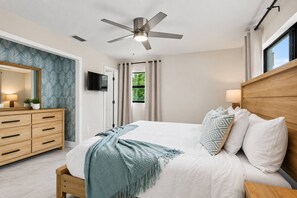 Een slaapkamer met een bed, nachtkastje, televisie, plafondventilator en een raam.