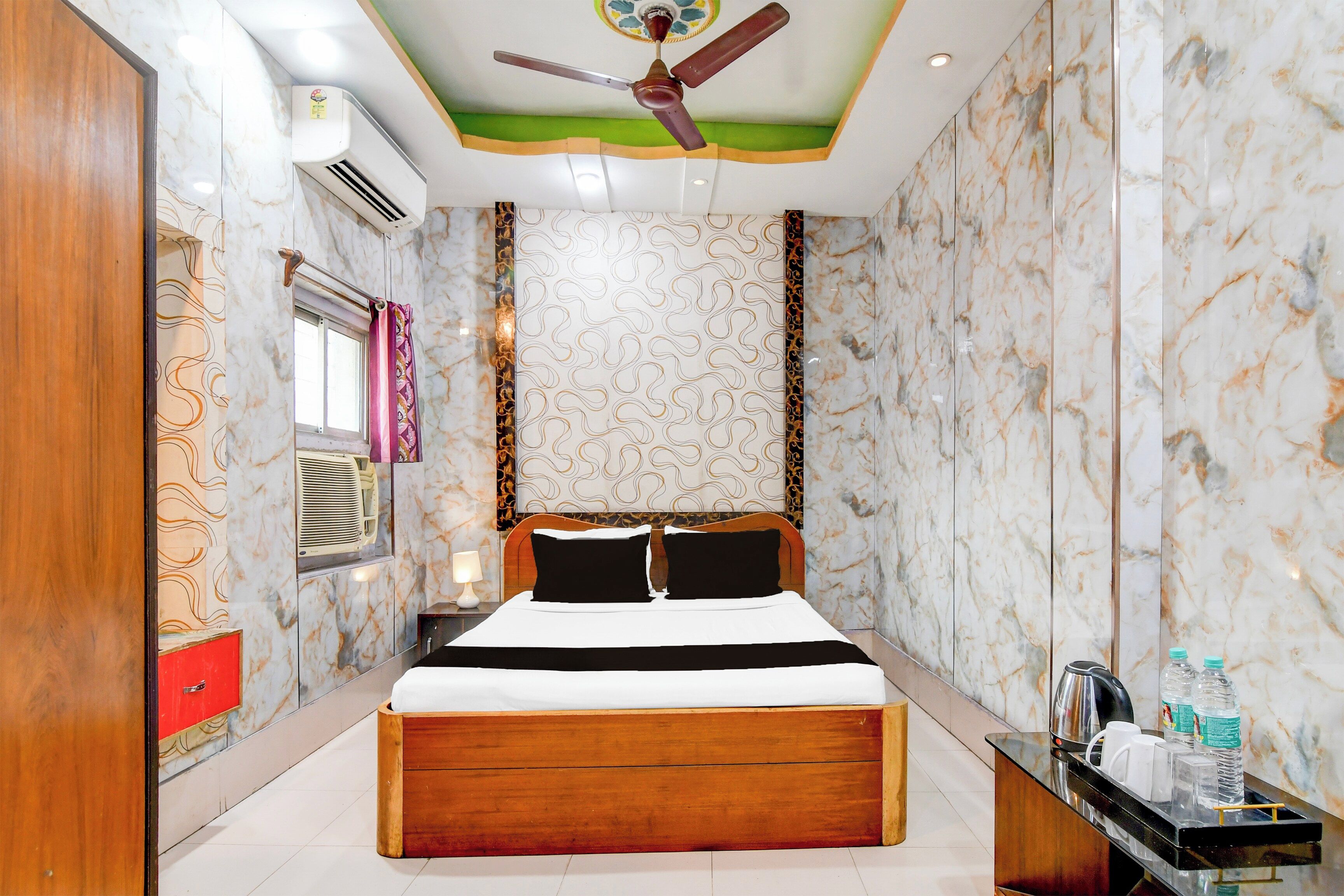 A hotel room with a bed, a ceiling fan, an air conditioning unit, a bedside table with a lamp, and a minibar.