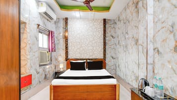A hotel room with a bed, a ceiling fan, an air conditioning unit, a bedside table with a lamp, and a minibar.