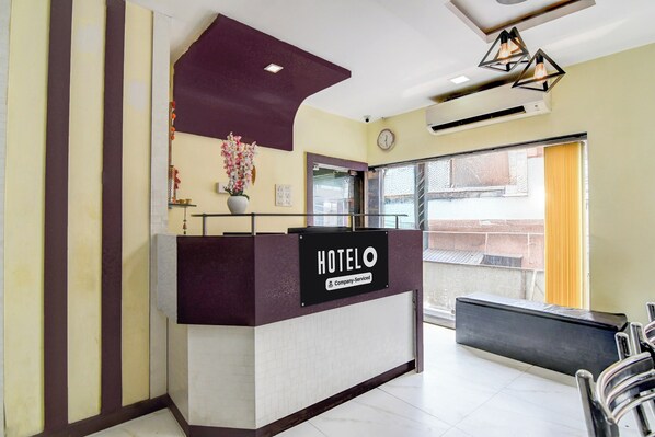 A hotel reception desk with a sign indicating it is company-serviced. There is a clock on the wall and a window with curtains.