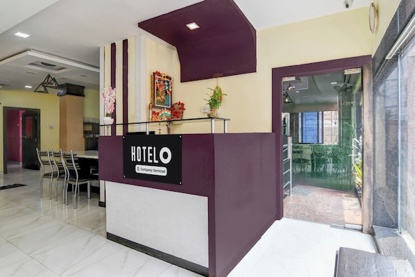 A hotel reception desk with a sign indicating it's company-serviced. There are chairs and a table in the background, and a door leading to another room.
