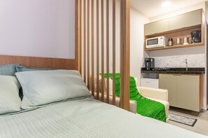 A modern hotel room with a bed, a green chair, a wooden partition, a kitchenette with a microwave, coffee maker, and a small refrigerator. - Smart Flat Premium | Alexa | Porto City Center | 210 (Porto de Galinhas)