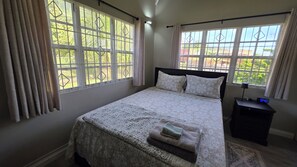 Ein Schlafzimmer mit einem Bett, Vorhängen, einem Nachttisch, einer digitalen Uhr und Blick auf Grünfläche.