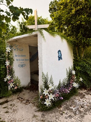 A small, rustic structure with a cross on top, surrounded by greenery and flowers.