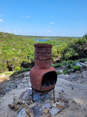 A rustic outdoor setting with a large, weathered pot on a stand, surrounded by natural terrain and a distant body of water.