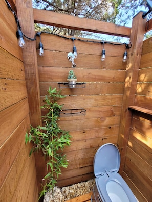 A wooden outdoor toilet with a metal shelf above it, a hanging light fixture, and a potted plant beside the toilet.