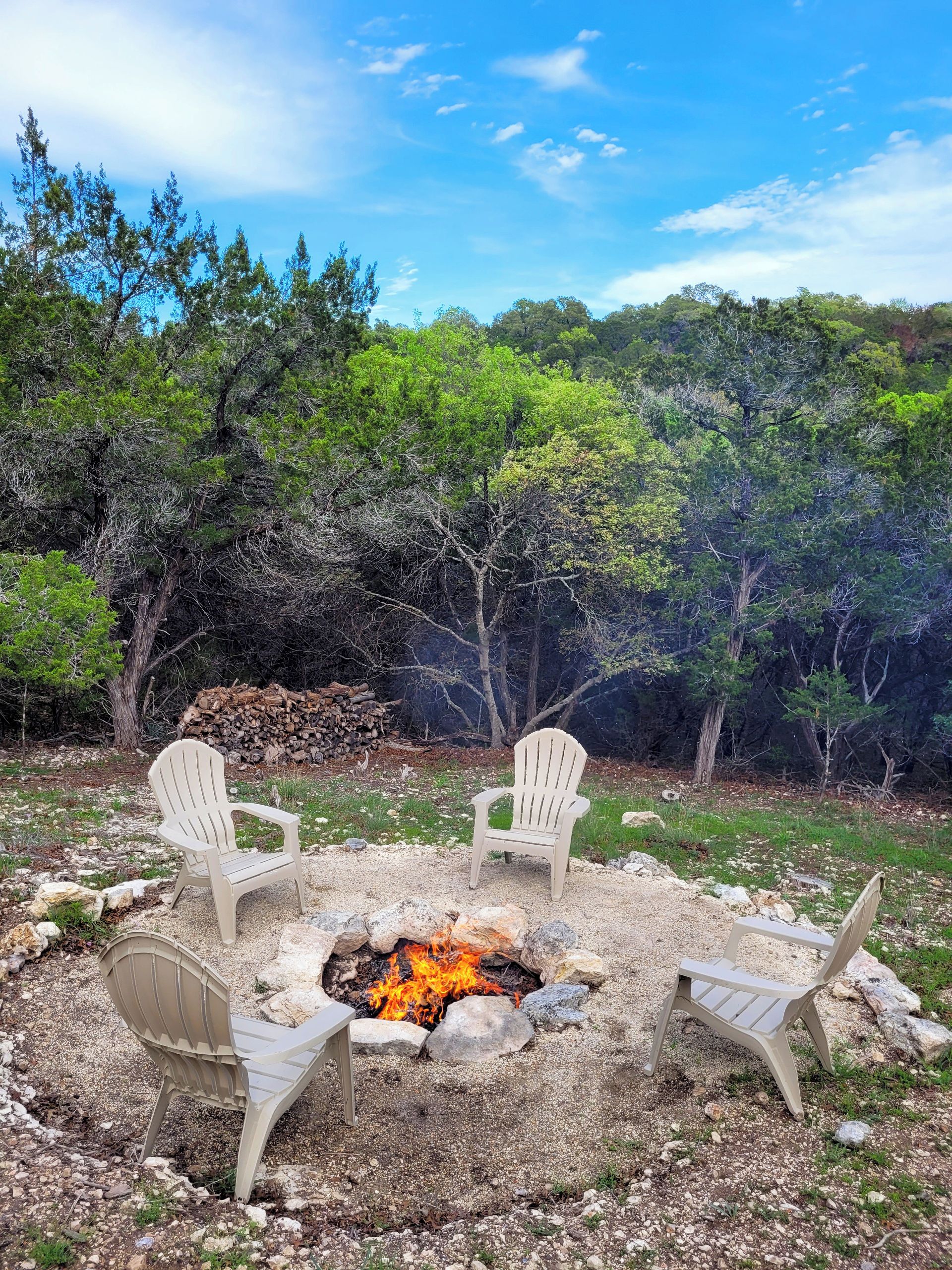 Un espacio al aire libre con sillas Adirondack alrededor de un brasero, en un entorno natural con árboles y cielo despejado.