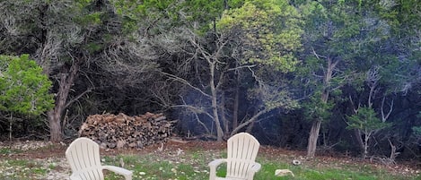 Un espacio al aire libre con sillas Adirondack alrededor de un brasero, en un entorno natural con árboles y cielo despejado.