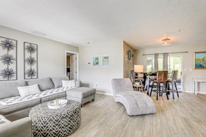 A living room with a grey sofa, a patterned ottoman, a grey armchair, and a dining area with a table and chairs. - 2BR Pet-Friendly – Walk or Bike to Manasota Beach (Englewood)