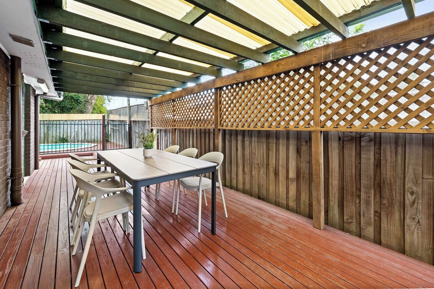 An outdoor patio with a wooden deck, a table with chairs, and a pool in the background.