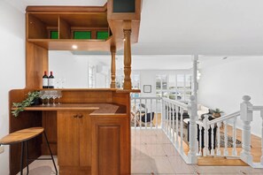 Una zona de bar con madera, botellas de vino y copas, un taburete y una escalera con barandillas blancas que sube a un nivel superior.