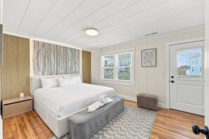A bedroom with a bed, bedside table, bench, and windows. - Elvi's Beach B | 3BR Coastal Getaway (North Myrtle Beach)
