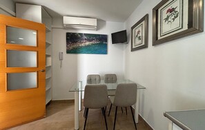 Ein Zimmer mit einem Glastisch, vier Stühlen, einer Klimaanlage, einem Wandfernseher und gerahmten Kunstwerken an der Wand.