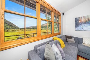 A room with a large window overlooking a green landscape, a grey sofa with cushions, and a painting on the wall. - New! Ski Days + Sunset Après | 3BR Mountain Retreat | Walk to Ski + Garage (Telluride)