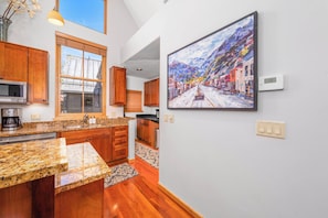 A modern kitchen with wooden cabinets, granite countertops, a large window, and a painting of a street scene on the wall. - New! Ski Days + Sunset Après | 3BR Mountain Retreat | Walk to Ski + Garage (Telluride)