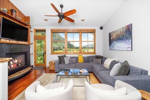 A living room with a fireplace, a large window with a view of a forest, a ceiling fan, and a painting on the wall. - New! Ski Days + Sunset Après | 3BR Mountain Retreat | Walk to Ski + Garage (Telluride)