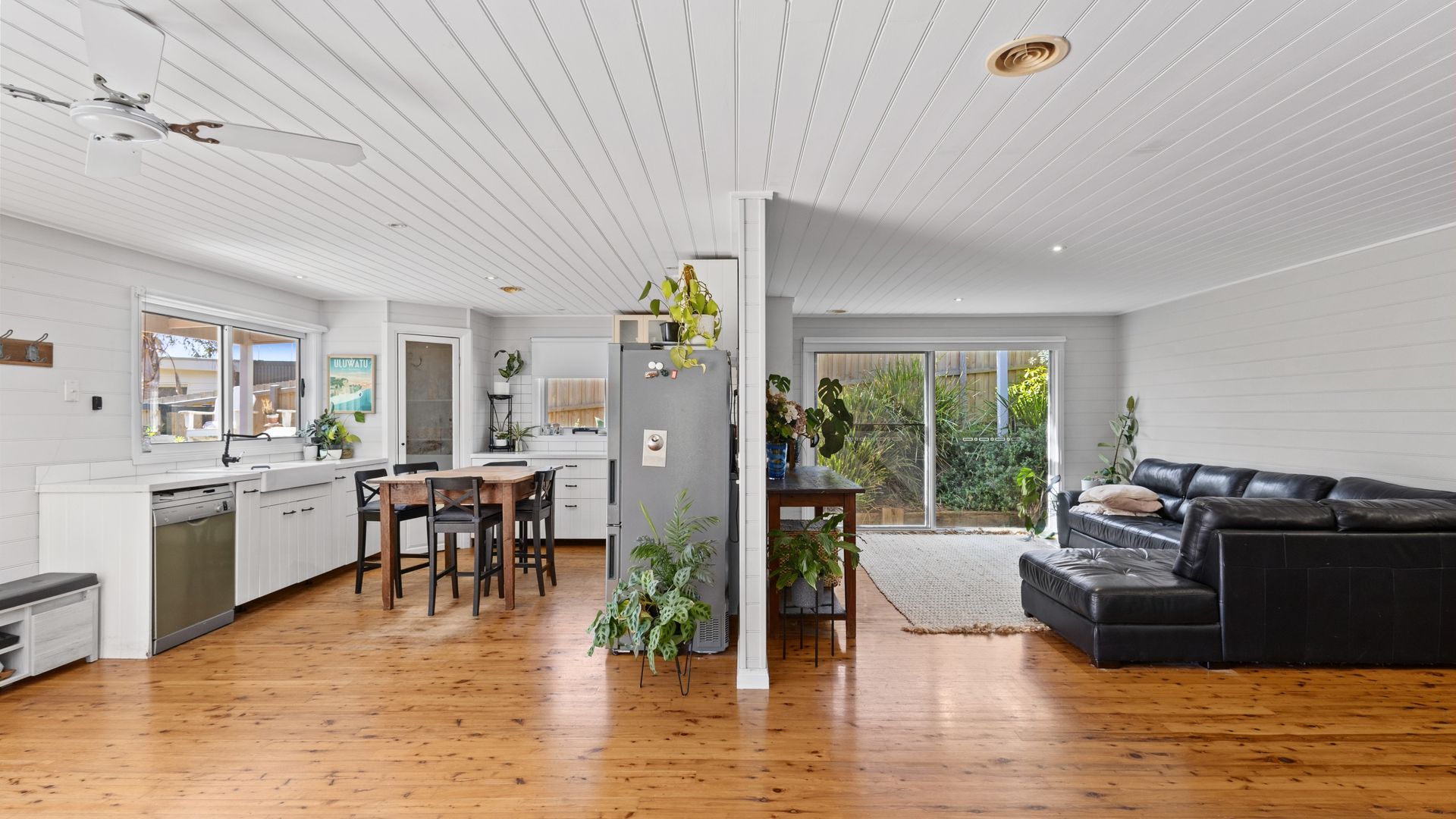 A spacious living area with a wooden floor, a black leather sofa, and a kitchen with white cabinetry.