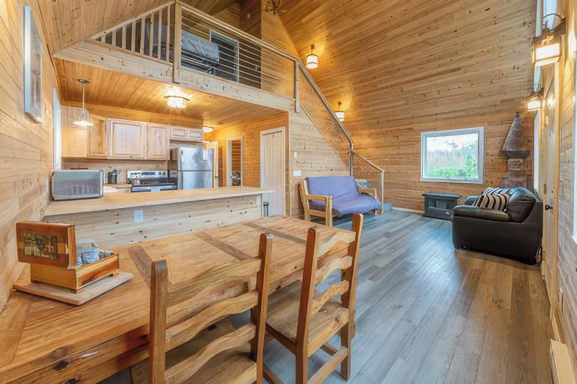 A wooden cabin interior with a dining area, kitchen, and a staircase leading to an upper level.
