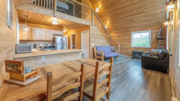 A wooden cabin interior with a dining area, kitchen, and a staircase leading to an upper level.