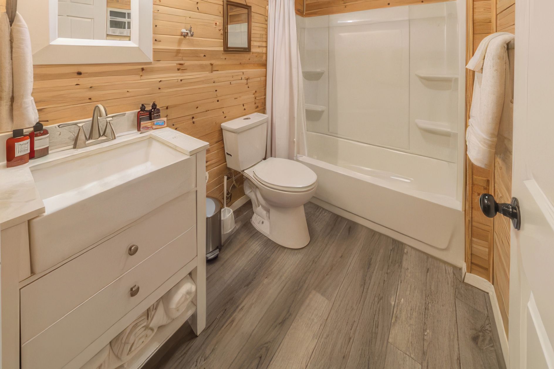 Un baño con pared de madera, inodoro blanco, bañera y lavamanos con artículos de aseo.