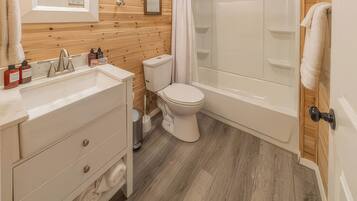 Un baño con pared de madera, inodoro blanco, bañera y lavamanos con artículos de aseo.