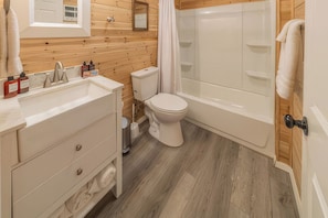 A bathroom with a wooden wall, a white toilet, a bathtub, and a sink with toiletries. - Gorgeous 2-bedroom cabin in charming Big Lake perfect for relaxing getaway (Big Lake)