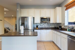 A modern kitchen with white cabinets, stainless steel appliances, a tiled backsplash, and a window with blinds. - The Lindenthal | Family Friendly 5BR | Fenced Yard | Sleeps 10 (Tucson)