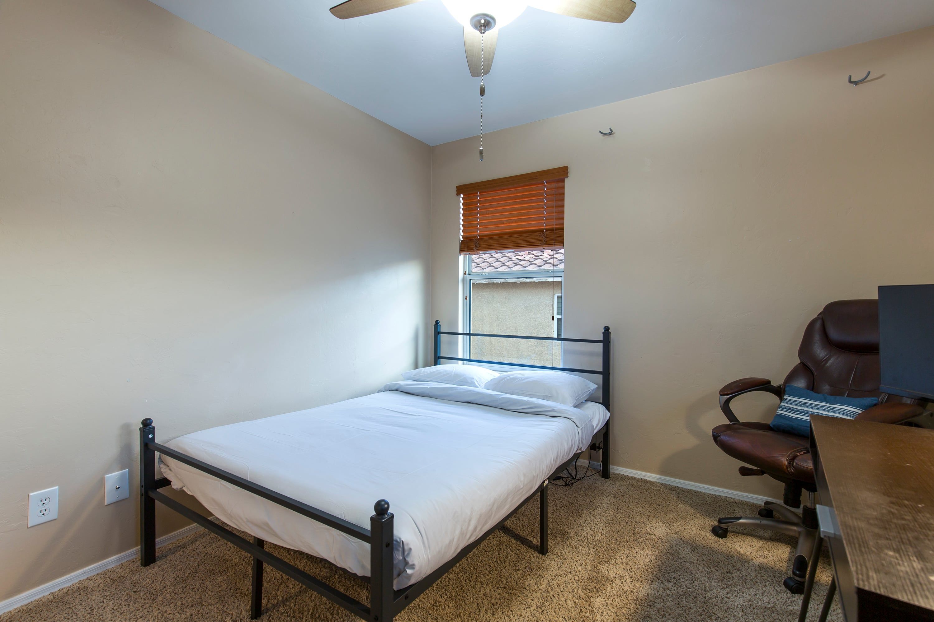 Une chambre avec un lit simple, un bureau avec un ordinateur, une chaise, un ventilateur de plafond et une fenêtre avec des stores.
