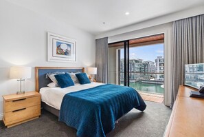 A hotel room with a large bed, a nightstand, a desk, a window with a view of buildings and water, and a framed picture on the wall. - Rare 2 Bed 2 Bath Corner Apartment at Latitude 37 (Auckland City)