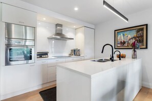 A modern kitchen with stainless steel appliances, a large island with a sink, and a painting on the wall. - Rare 2 Bed 2 Bath Corner Apartment at Latitude 37 (Auckland City)