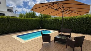 Ein Swimmingpool mit einer Holzterrasse, zwei Korbstühlen, einem Tisch und einem großen Sonnenschirm.