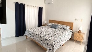 Ein Schlafzimmer mit Bett, Vorhängen, Deckenventilator, Klimaanlage, Nachttisch mit Lampe und an der Wand montiertem Fernseher.