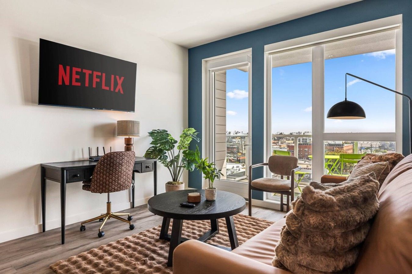 Ein modernes Wohnzimmer mit einem großen Fernseher, der 'NETFLIX' anzeigt, einem bequemen Sofa, einem Couchtisch und einem Blick auf die Stadt durch große Fenster.