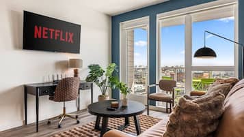 Ein modernes Wohnzimmer mit einem großen Fernseher, der 'NETFLIX' anzeigt, einem bequemen Sofa, einem Couchtisch und einem Blick auf die Stadt durch große Fenster.