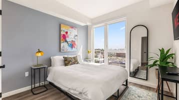 Un dormitorio moderno con una cama grande, una lámpara de noche, un cuadro y vistas a través de un ventanal.