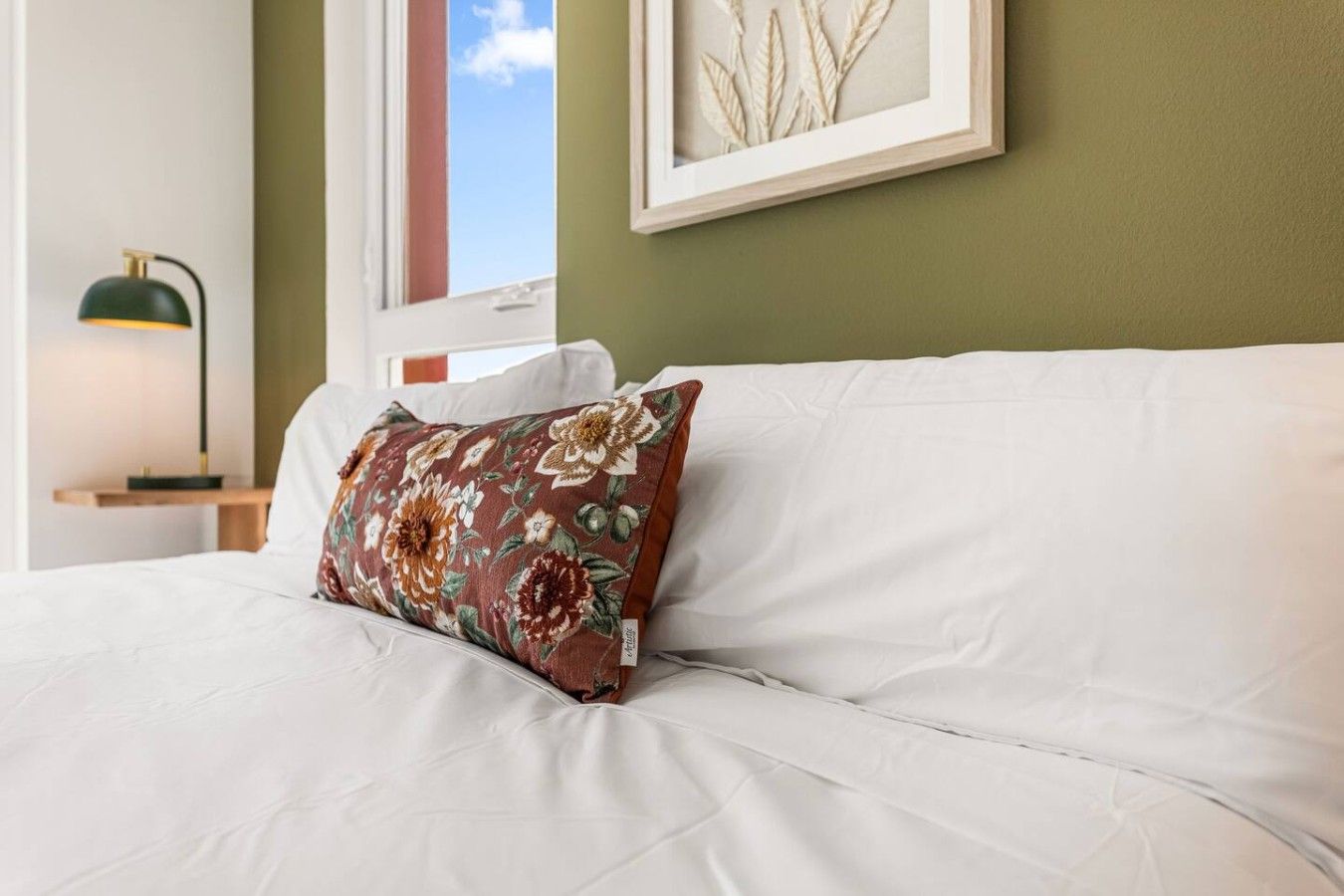 A bed with a white mattress and a floral pillow, a bedside table with a lamp, and a framed artwork on the wall.