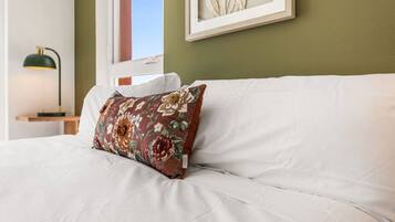 A bed with a white mattress and a floral pillow, a bedside table with a lamp, and a framed artwork on the wall.
