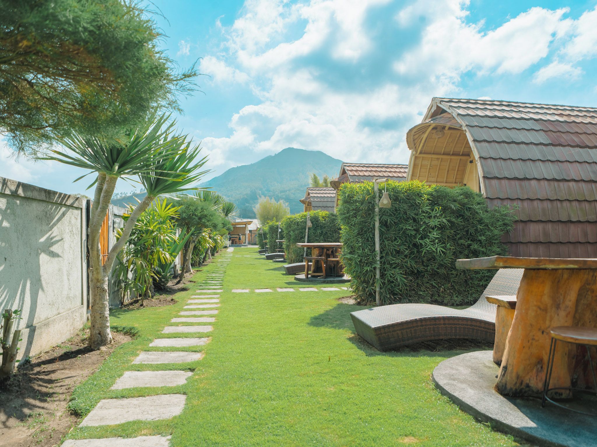 Un sendero conduce a un edificio con techo tradicional, rodeado de abundante vegetación y una montaña al fondo.
