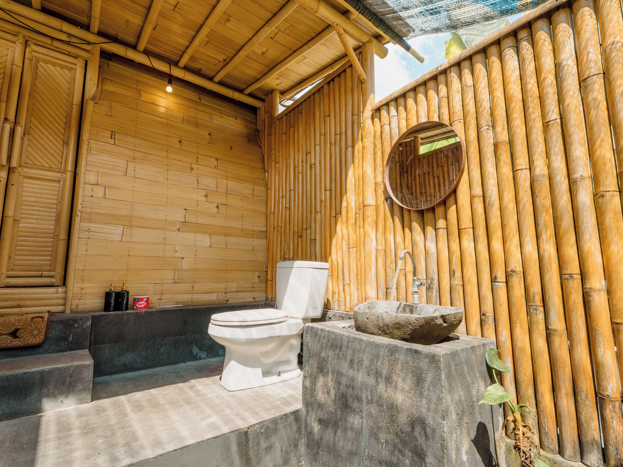 Une salle de bain aux murs en bambou, équipée de toilettes, d’un lavabo en pierre et d’une petite fenêtre.