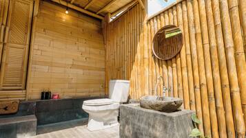 Une salle de bain aux murs en bambou, équipée de toilettes, d’un lavabo en pierre et d’une petite fenêtre.
