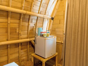 A compact room with bamboo walls, a small white refrigerator, a blue kettle, and a woven basket.