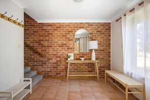 A room with a brick wall, a wooden bench, a small table with a lamp, and a staircase.