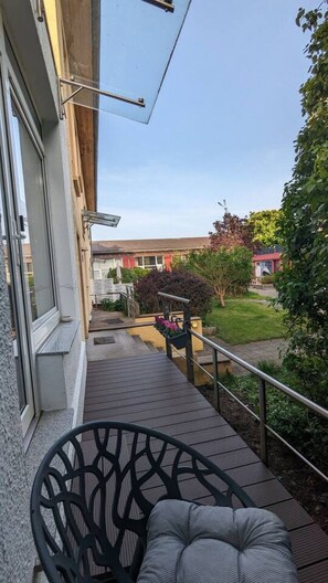 Ein Balkon mit Stuhl und Tisch, eine Holzterrasse und Blick auf eine Wohngegend mit Häusern und Grünflächen.