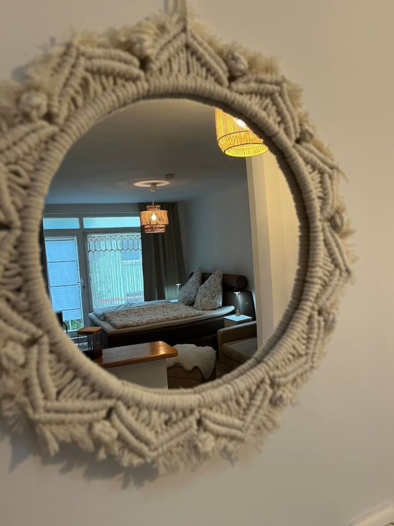 Een ronde spiegel met een reliëfomlijsting die een slaapkamer weerspiegelt, met een bed, nachtkastje en hanglamp.
