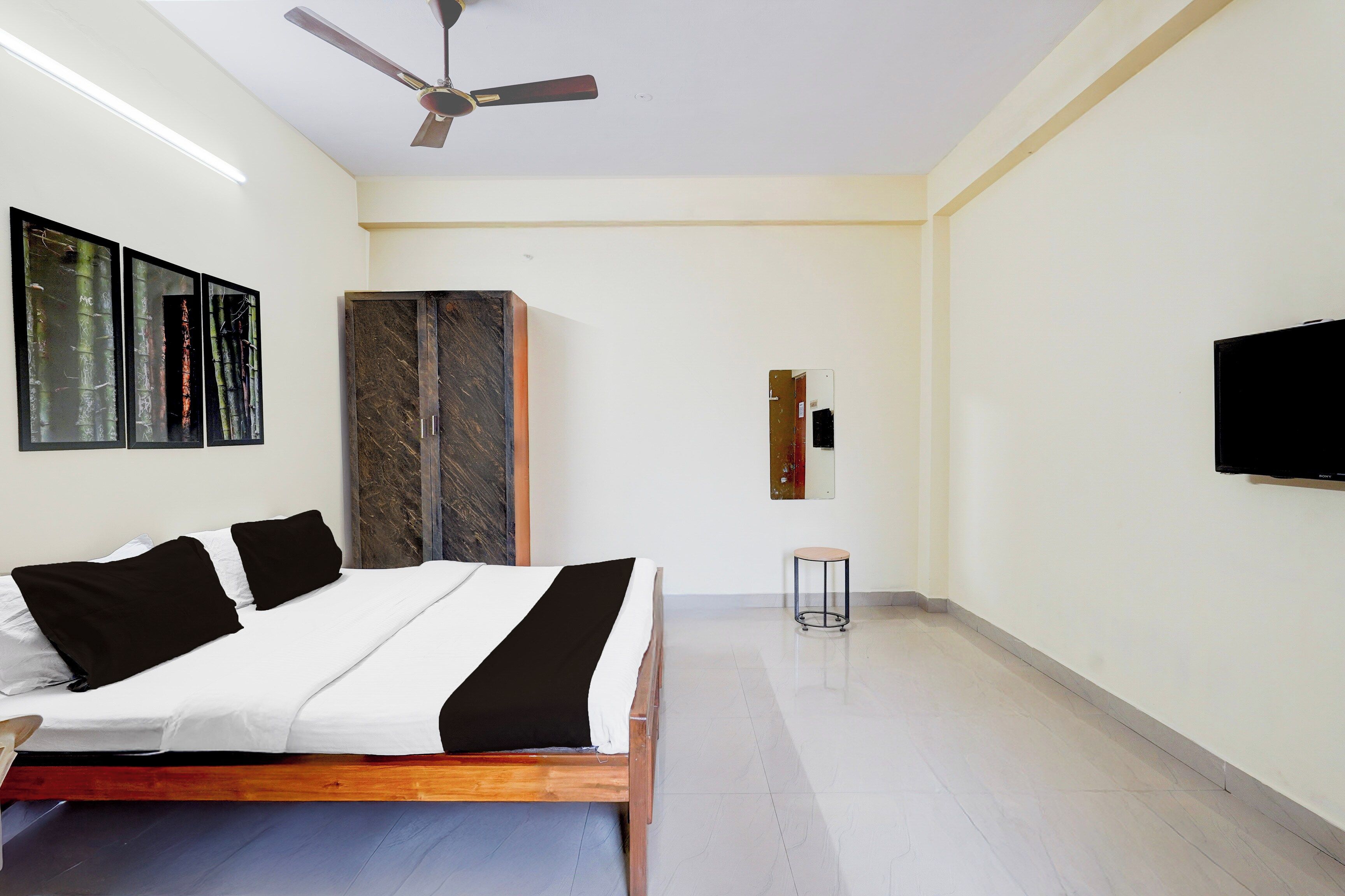 A bedroom with a bed, a wardrobe, a ceiling fan, a small stool, and a flat-screen TV mounted on the wall.