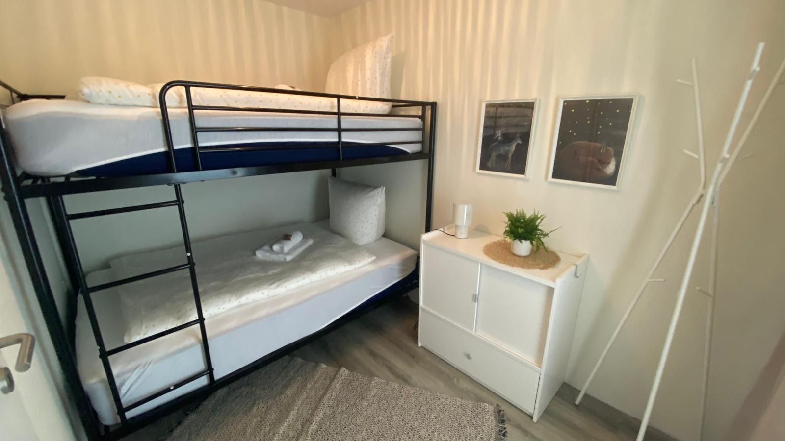 Ein Etagenbett mit Schreibtisch und einer kleinen Pflanze, ein weißer Schrank und zwei gerahmte Bilder an der Wand.